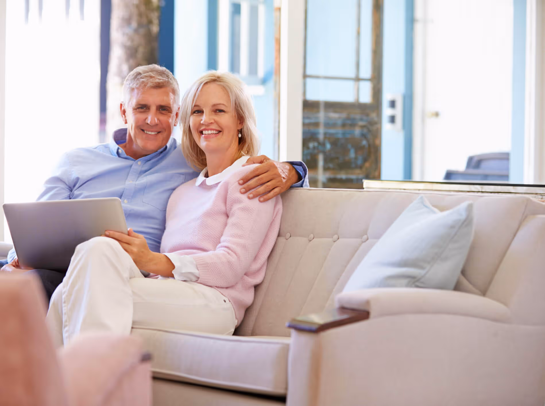Smiling middle-aged couple sitting closely on a beige couch, holding a laptop in a bright living room.