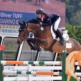 Equestrian rider wearing black helmet and jacket jumping a brown and white horse over a green and white hurdle.