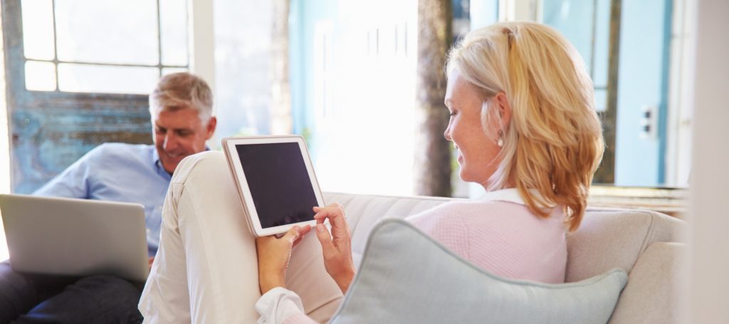 Man and women sat on a sofa looking at a laptop and a tablet. 