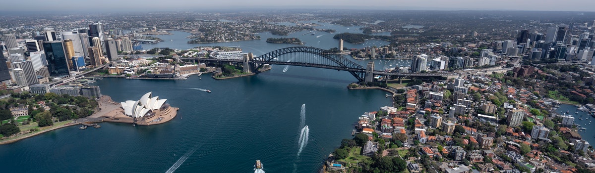 An high drone photo of Sydney with a bridge and the Opera house in view on a sunny day. 