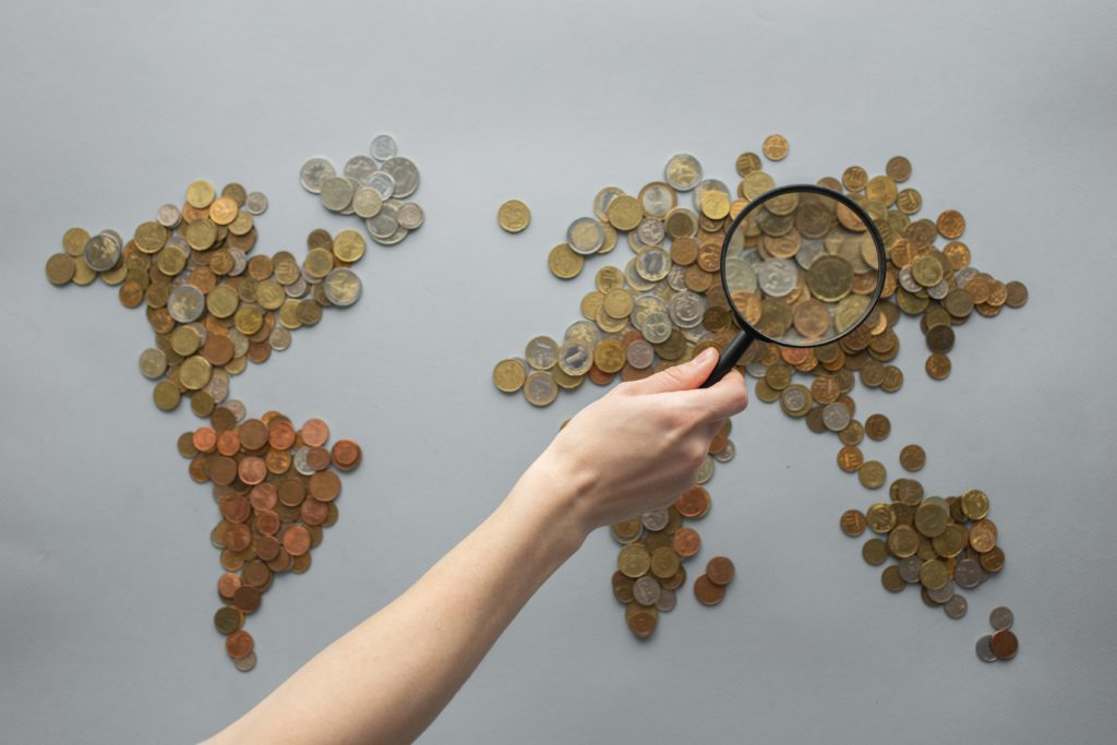 world map created by using coins with a person holding a magnifying glass over a certain area.