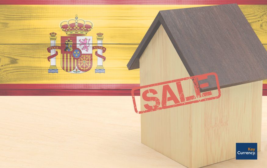 Spanish flag in the background with a wooden house placed on a table at the front. There is also a read Sale sign stamped on the house. 
