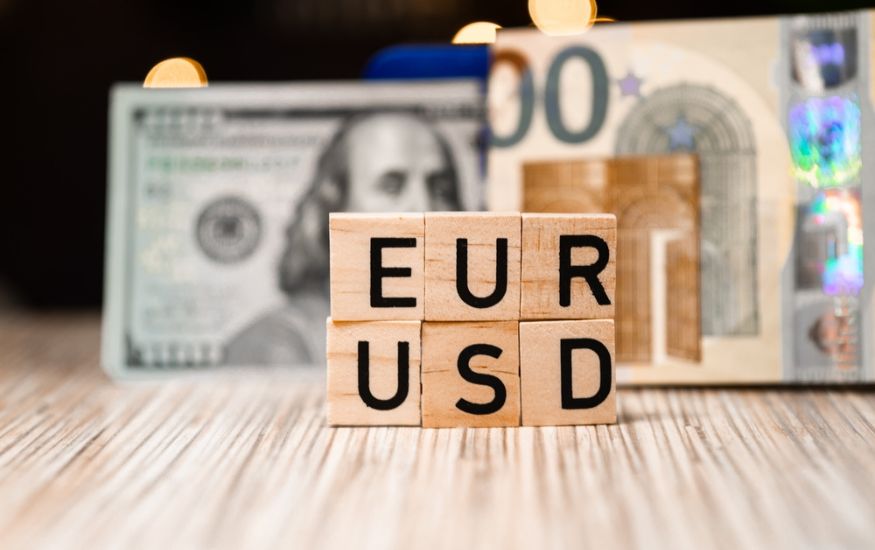 wooden blocks that spell out EUR and USD stacked on top of one another. In the background there is a dollar note and a euro note. The image depicts a euro to usd money transfer. 